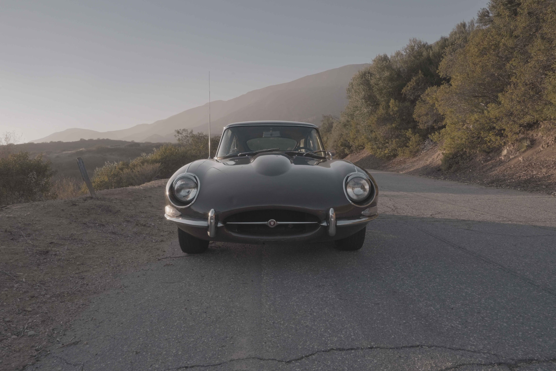Head-on view of a black 1968 Jaguar E-Type on a canyon road at dawn