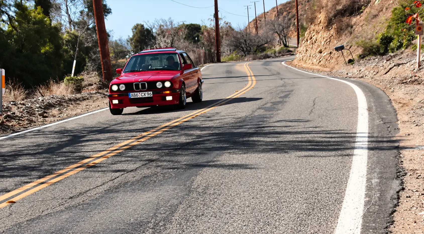 Red BMW E30 rounding a sunlit curve on a mountain road